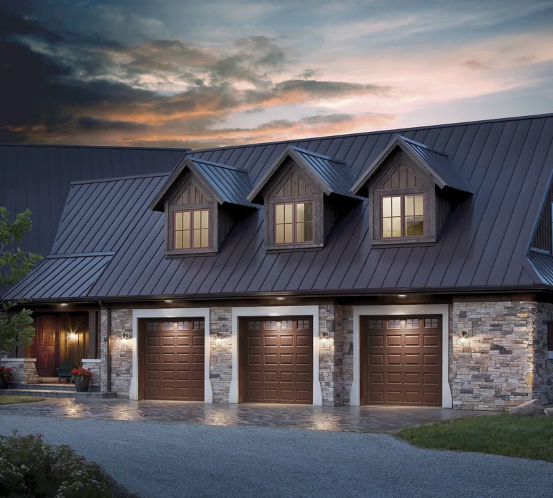 Clopay Classic Steel wood-tone carriage garage doors on a metal-roof home at dusk